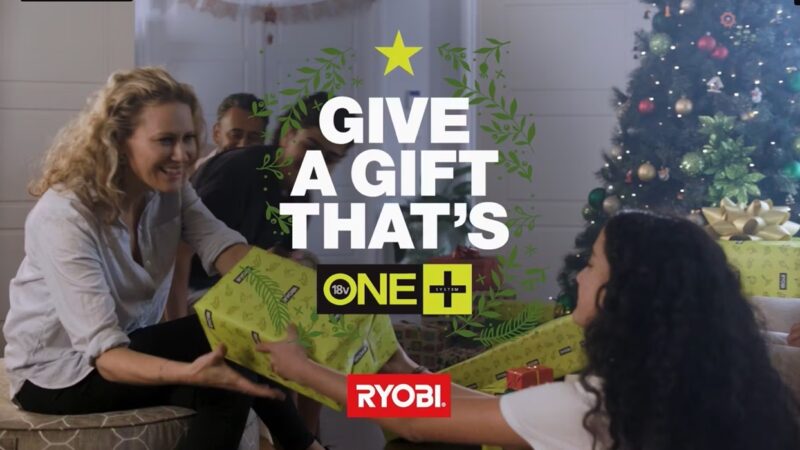 woman handing girl gift in front of Christmas tree with family. Text on screen give a gift that's one+ Ryobi
